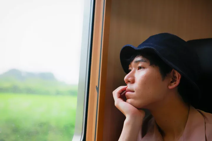 Молодой японец в панаме задумчиво смотрит в окно поезда.
